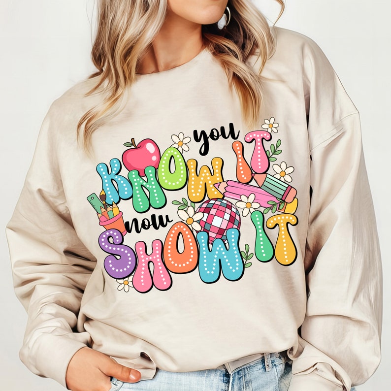 K&ouml;nnte beinhalten: Ein beiger Pullover mit einem bunten Grafikdesign, auf dem "You Know It Now Show It" mit einem roten Apfel, Bleistiften und Blumen steht.