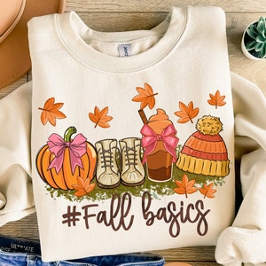 May include: A beige sweatshirt with a graphic design of a pumpkin, boots, a coffee drink, and a beanie hat. The graphic is surrounded by fall leaves and the text "#Fall basics".