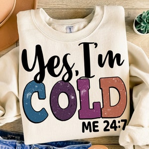 以下が含まれることがあります： クリーム色のスウェットシャツに、「Yes, I'm COLD ME 24:7」という黒い文字とカラフルな文字が書かれています。文字はレトロなスタイルで、使い古したような風合いになっています。
