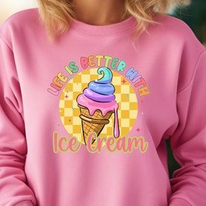 Puede incluir: Sudadera rosa con un gr&aacute;fico de un cono de helado colorido en un cono de waffle. El texto "Life is better with ice cream" est&aacute; impreso en la sudadera.