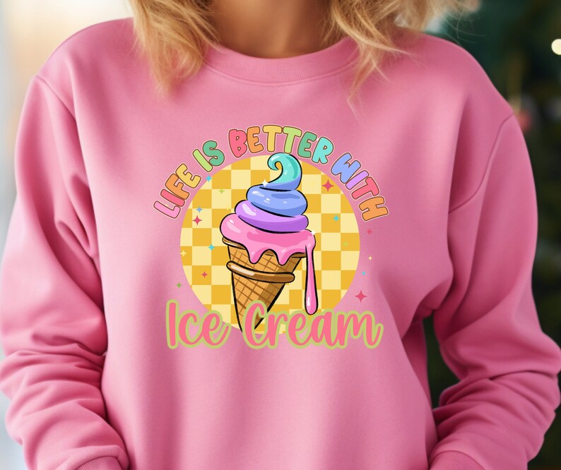 Puede incluir: Sudadera rosa con un gr&aacute;fico de un cono de helado colorido en un cono de waffle. El texto "Life is better with ice cream" est&aacute; impreso en la sudadera.