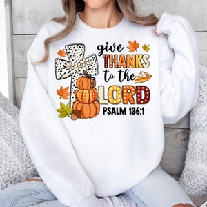 May include: White sweatshirt with a graphic of a cross, pumpkins, and a pie. The text reads "give thanks to the Lord Psalm 136:1".