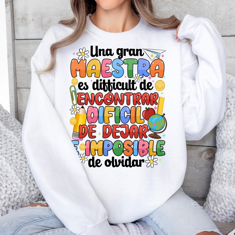 Op de afbeelding: Wit sweatshirt met een kleurrijk grafisch ontwerp met de tekst "Una gran maestra es difficult de encontrar dificil de dejar imposible de olvidar". De grafiek bevat een potlood, een liniaal, een aardbol, een papieren vliegtuig en bloemen.