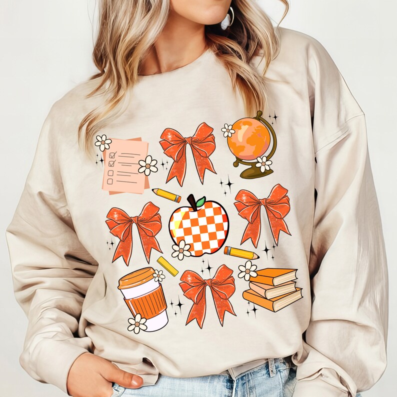 May include: A beige sweatshirt with an orange and white design featuring a checkered apple, bows, a globe, books, a pencil, a coffee cup, and a checklist.