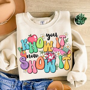 K&ouml;nnte beinhalten: Ein beiger Pullover mit einem bunten Grafikdesign, auf dem "You Know It Now Show It" mit einem roten Apfel, Bleistiften und Blumen steht.