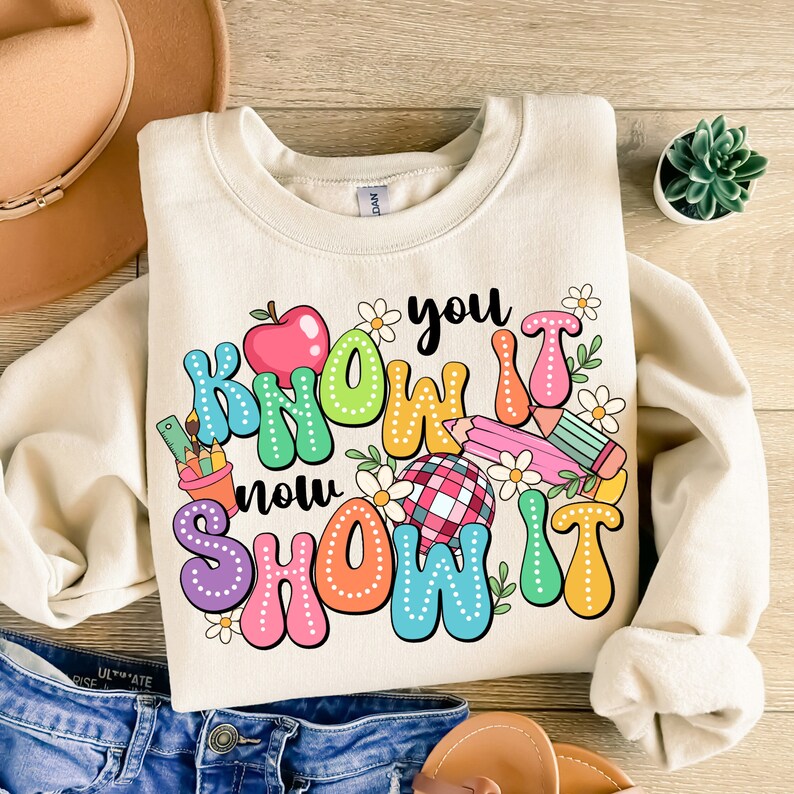 K&ouml;nnte beinhalten: Ein beiger Pullover mit einem bunten Grafikdesign, auf dem "You Know It Now Show It" mit einem roten Apfel, Bleistiften und Blumen steht.