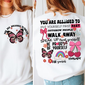 May include: White sweatshirt with a pink butterfly and the text "You are allowed to thrive" on the front. The back of the sweatshirt has the text "You are allowed to put yourself first rest outgrow people walk away speak up treat yourself be proud of yourself change for yourself" with pink butterflies, cherries, a rainbow, and flowers.
