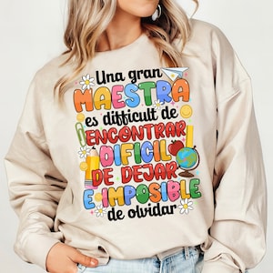 Op de afbeelding: Een beige sweatshirt met een kleurrijk grafisch ontwerp met de tekst "Una gran MAESTRA es difficult de ENCONTRAR DIFCIL DE DEJAR E IMPOSIBLE de olvidar".