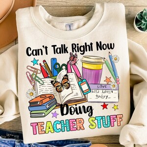 May include: A beige sweatshirt with the text "Can't Talk Right Now Doing Teacher Stuff" in colorful letters. The design includes a book, coffee cup, pencils, scissors, paper clips, and a butterfly.