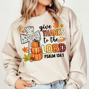 May include: A beige sweatshirt with a graphic design featuring a black and white cross with white daisies, a stack of pumpkins, and the text "give THANKS to the LORD PSALM 136:1".