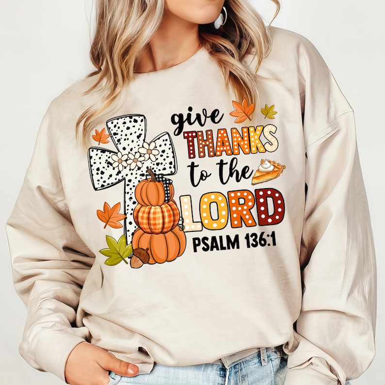 May include: A beige sweatshirt with a graphic design featuring a black and white cross with white daisies, a stack of pumpkins, and the text "give THANKS to the LORD PSALM 136:1".