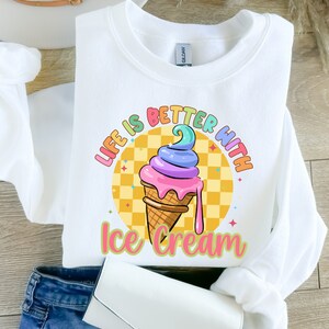 Puede incluir: Sudadera blanca con un gr&aacute;fico de cono de helado colorido y el texto "Life is better with ice cream".