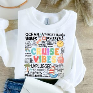 Peut inclure: Sweat-shirt blanc avec un motif graphique color&eacute; comportant les mots "Ocean Waves", "Cruise Vibes", "Unplugged", "Seashells", "Memories", et "Paradise Awaits". Le graphique comprend &eacute;galement des images d'un bateau de croisi&egrave;re, d'une valise, d'un palmier et d'un coquillage.