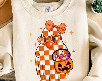 Pode incluir: Uma camisa de moletom branca com um fantasma de desenho animado fofo usando óculos de sol xadrez laranja e branco, um laço rosa e segurando um balde de abóbora cheio de doces. O fantasma tem um nariz vermelho e um sorriso amigável.