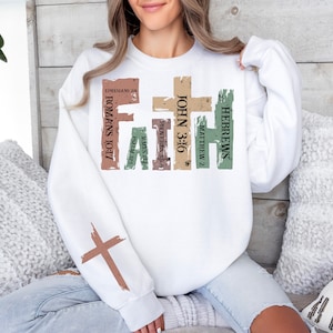 Puede incluir: Sudadera blanca con un dise&ntilde;o de cruz desgastada y la palabra "FAITH" en letras grandes. La cruz est&aacute; formada por vers&iacute;culos b&iacute;blicos, incluyendo "Hebreos", "Mateo 2", "Juan 3:16", "Proverbios 3:5-6", "Santiago 2:1", "Salmo 37:5", "Romanos 10:17" y "Efesios 2:8".