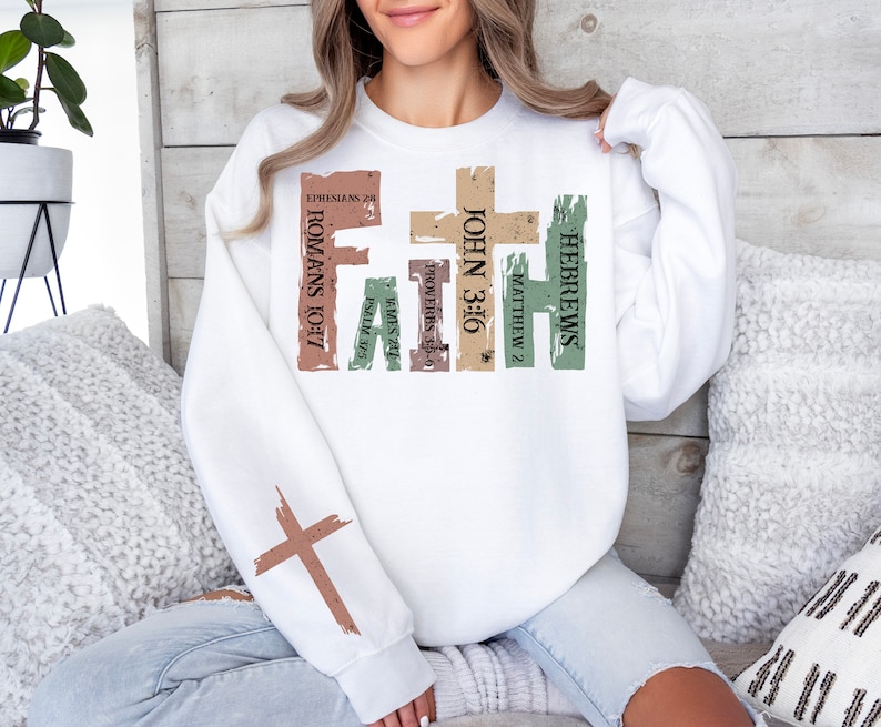 Puede incluir: Sudadera blanca con un dise&ntilde;o de cruz desgastada y la palabra "FAITH" en letras grandes. La cruz est&aacute; formada por vers&iacute;culos b&iacute;blicos, incluyendo "Hebreos", "Mateo 2", "Juan 3:16", "Proverbios 3:5-6", "Santiago 2:1", "Salmo 37:5", "Romanos 10:17" y "Efesios 2:8".
