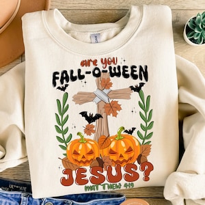 Könnte beinhalten: Ein weißer Pullover mit einem grafischen Design, das ein Kreuz mit Kürbissen, Fledermäusen und Blättern zeigt. Der Text lautet "Are you Fall-o-ween Jesus? Matthew 4:19".