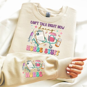 Puede incluir: Sudadera beige con un gráfico colorido que dice "Can't Talk Right Now Doing Nurse Stuff". El diseño incluye una jeringa, un portapapeles, una taza de café, un frasco de medicamentos y un estetoscopio. La manga también tiene un gráfico de "Nurse".