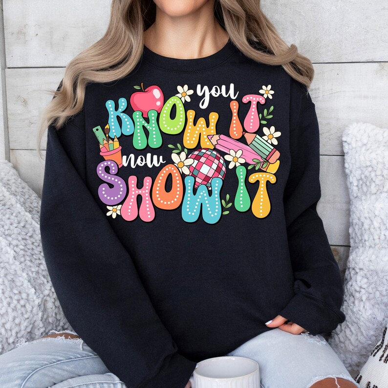 K&ouml;nnte beinhalten: Schwarzer Pullover mit einer bunten Grafik, auf der "Know it now show it" mit einem roten Apfel, Bleistiften und Blumen steht.