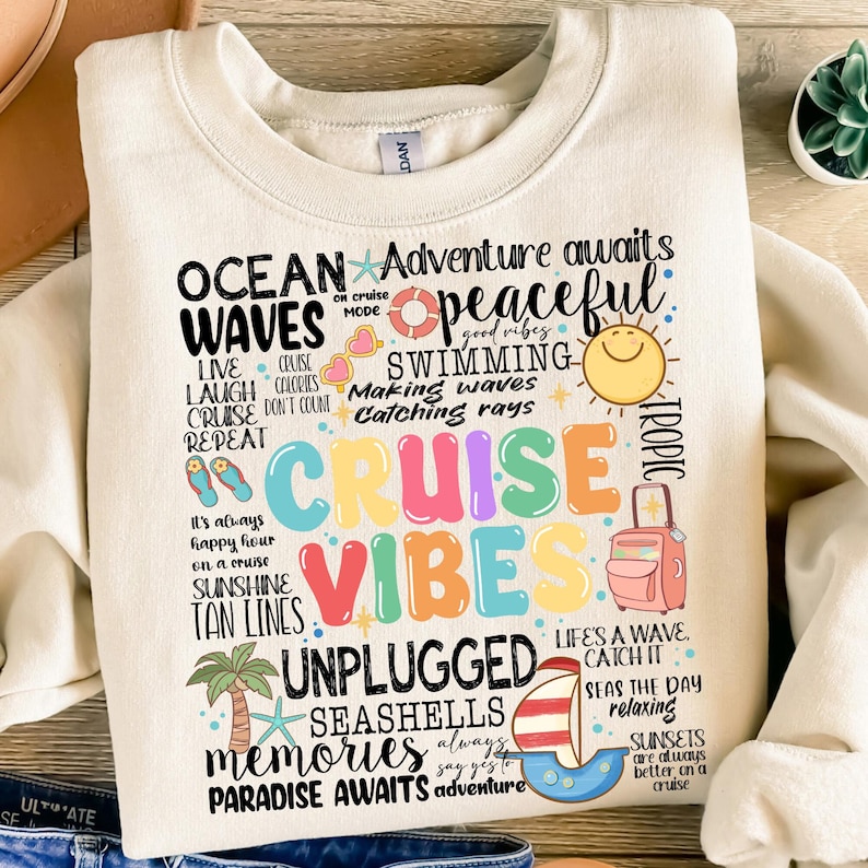 Peut inclure: Un sweat-shirt blanc avec un motif graphique color&eacute; comportant le texte "Ocean Waves", "Cruise Vibes", "Unplugged", "Seashells", "Memories", "Paradise Awaits", et "Adventure Awaits". Le graphique comprend &eacute;galement des images d'un soleil, d'une valise, d'un palmier et d'un voilier.