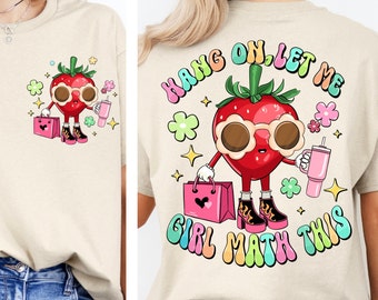 May include: A beige t-shirt with a cartoon strawberry wearing sunglasses and holding shopping bags. The text "Hang on, let me girl math this" is printed in rainbow colors on the shirt.