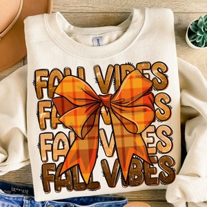 Peut inclure: Un sweat-shirt crème avec le texte "Fall Vibes" répété quatre fois dans une police d'écriture vieillie. Un grand nœud en plaid orange et brun est centré sur le sweat-shirt.