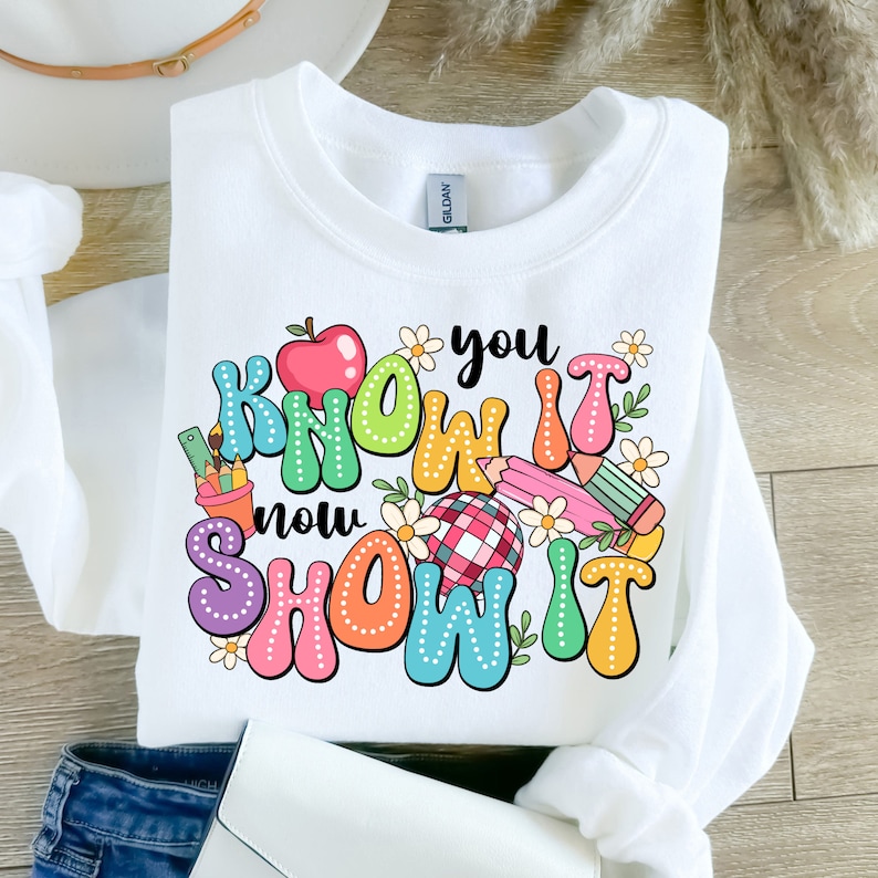 K&ouml;nnte beinhalten: Wei&szlig;er Pullover mit einem bunten Grafikdesign, auf dem "You know it now show it" mit einem roten Apfel, Bleistiften und Blumen steht.