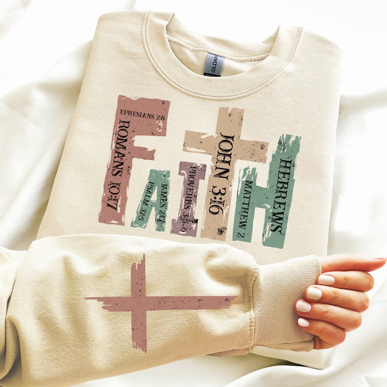 Puede incluir: Una sudadera beige con un dise&ntilde;o gr&aacute;fico que presenta la palabra "FAITH" en una fuente colorida y desgastada. El dise&ntilde;o incluye varios vers&iacute;culos b&iacute;blicos, entre ellos "Juan 3:16", "Romanos 10:17" y "Hebreos".