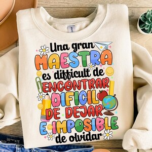 Op de afbeelding: Een beige sweatshirt met een kleurrijk grafisch ontwerp met de tekst "Una gran maestra es difficult de encontrar, dificil de dejar, e imposible de olvidar". De grafiek bevat een potlood, een liniaal, een wereldbol, een bloem en een rode appel.