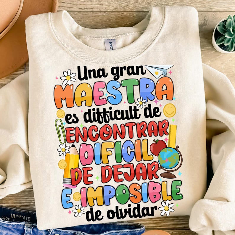 Op de afbeelding: Een beige sweatshirt met een kleurrijk grafisch ontwerp met de tekst "Una gran maestra es difficult de encontrar, dificil de dejar, e imposible de olvidar". De grafiek bevat een potlood, een liniaal, een wereldbol, een bloem en een rode appel.