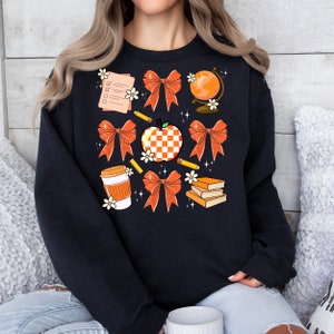May include: Black sweatshirt with a graphic design of orange and white bows, a checkered apple, a globe, pencils, a coffee cup, and books. The design is surrounded by white flowers.