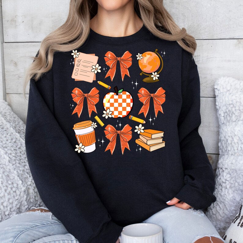 May include: Black sweatshirt with a graphic design of orange and white bows, a checkered apple, a globe, pencils, a coffee cup, and books. The design is surrounded by white flowers.
