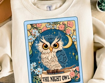 Op de afbeelding: Een crèmekleurig sweatshirt met een vintage-illustratie van een uil met blauwe en bruine veren. De uil zit op een tak met een halve maan en sterren op de achtergrond. De tekst "THE NIGHT OWL" is onder de uil gedrukt.