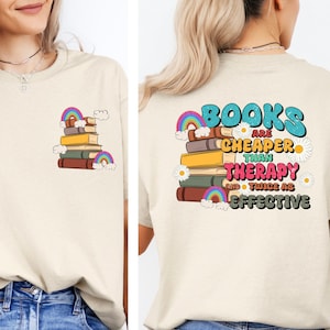 May include: A beige t-shirt with a graphic design of a stack of books with rainbows and the text "Books are cheaper than therapy and twice as effective".
