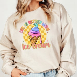 Puede incluir: Una sudadera beige con el texto "Life is better with ice cream" impreso en la parte delantera. El texto est&aacute; en colores arco&iacute;ris y hay un cono de helado de dibujos animados con chispas de arco&iacute;ris y un fondo amarillo.