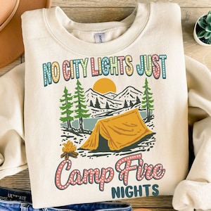 May include: A cream colored sweatshirt with the text "NO CITY LIGHTS JUST Camp Fire NIGHTS" in a retro style font. The text is surrounded by a graphic of a tent, a campfire, and mountains.