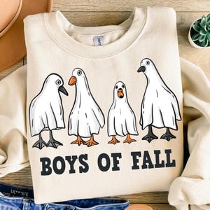 Pode incluir: Uma camisa de moletom cor creme com um gráfico de quatro gansos brancos vestindo fantasias de fantasmas. O texto "Boys of Fall" é impresso abaixo dos gansos.