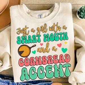 Puede incluir: Una sudadera beige con el texto "Just a girl with a smart mouth and a cornbread accent" en letras coloridas de estilo retro. Una sartén de dibujos animados con una rebanada de pan de maíz se representa junto al texto.