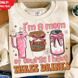 Op de afbeelding: Een beige sweatshirt met de tekst "i'm a mom of course i have THREE DRINKS" in bruin en roze letters. De tekst wordt omringd door drie illustraties van drankjes: een roze beker met een roze strik, een bruine koffiebeker met een roze strik en een roze blikje met een roze strik.