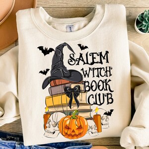 Może przedstawiać: Biała bluza z czarnym kapeluszem czarownicy, stos książek i dynią. Na bluzie nadrukowany jest tekst "Salem Witch Book Club".