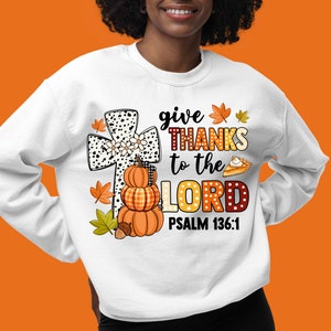 May include: White sweatshirt with a graphic design featuring a black and white cross with a stack of pumpkins, fall leaves, and a pie. The text reads "give THANKS to the LORD PSALM 136:1".