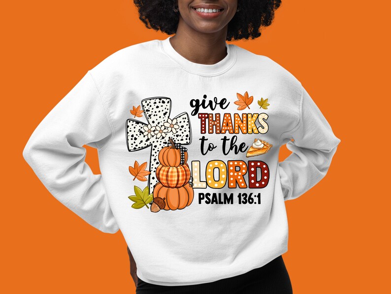 May include: White sweatshirt with a graphic design featuring a black and white cross with a stack of pumpkins, fall leaves, and a pie. The text reads "give THANKS to the LORD PSALM 136:1".