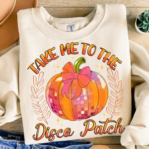 May include: A cream colored sweatshirt with the text "Take Me To The Disco Patch" in orange and pink. A disco ball pumpkin with a pink bow is in the center of the text.