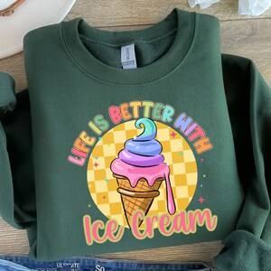 Puede incluir: Una sudadera verde oscuro con el texto "Life is better with ice cream" en colores arco&iacute;ris. Un cono de helado de dibujos animados con chispas de arco&iacute;ris est&aacute; en el centro del texto.