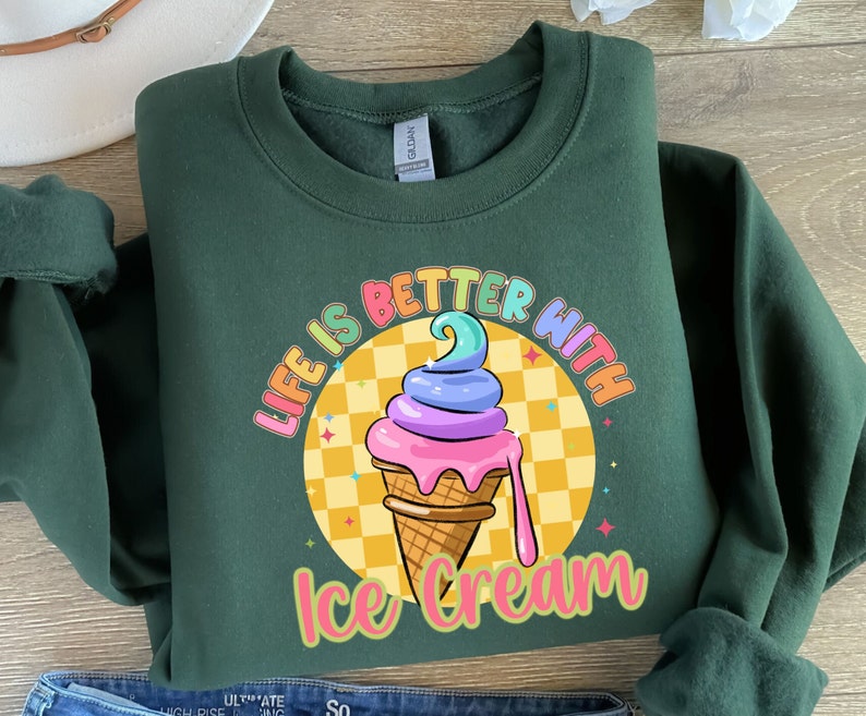 Puede incluir: Una sudadera verde oscuro con el texto "Life is better with ice cream" en colores arco&iacute;ris. Un cono de helado de dibujos animados con chispas de arco&iacute;ris est&aacute; en el centro del texto.
