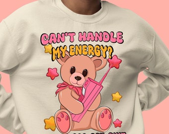 May include: A cream-colored sweatshirt with a pink teddy bear holding a pink phone. The text on the sweatshirt reads "Can't handle my energy? Dial: 1-800-EAT-SHIT".