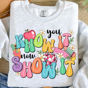 K&ouml;nnte beinhalten: Ein graues Sweatshirt mit einem bunten Grafikdesign, auf dem "Know it now show it" mit einem roten Apfel, Bleistiften und Blumen steht.
