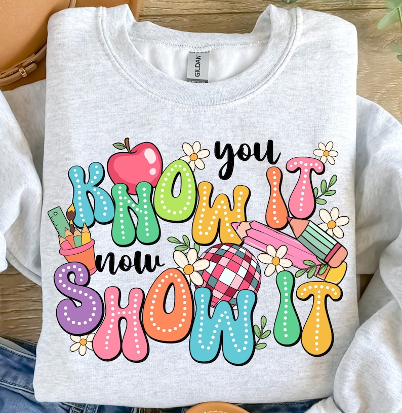 K&ouml;nnte beinhalten: Ein graues Sweatshirt mit einem bunten Grafikdesign, auf dem "Know it now show it" mit einem roten Apfel, Bleistiften und Blumen steht.