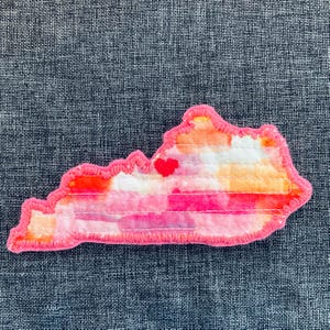 May include: A fabric patch in the shape of the state of Virginia, featuring a watercolor design in shades of pink, orange, and white. A small red heart is stitched on the patch. The patch is outlined with pink stitching.