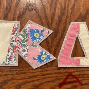 Może przedstawiać: Zbliżenie liter z tkaniny, które wymawiają słowo "K A". Litera "K" wykonana jest z kremowej tkaniny z kwiatowym wzorem i różowej tkaniny z niebieskimi kwiatami. Litera "A" jest różowa z białymi szwami.